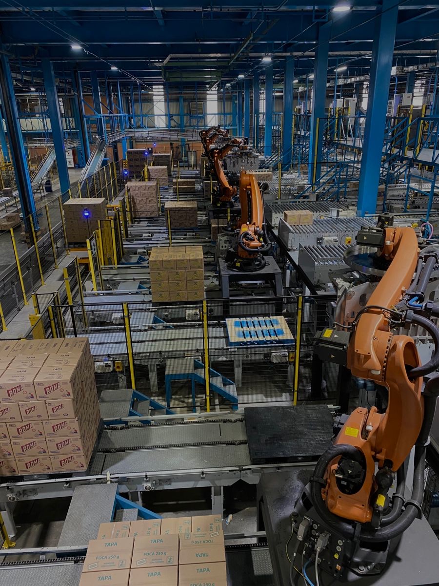 Vista elevada de una planta de manufactura donde varios brazos robóticos anaranjados paletizan cajas de cartón sobre cintas transportadoras.